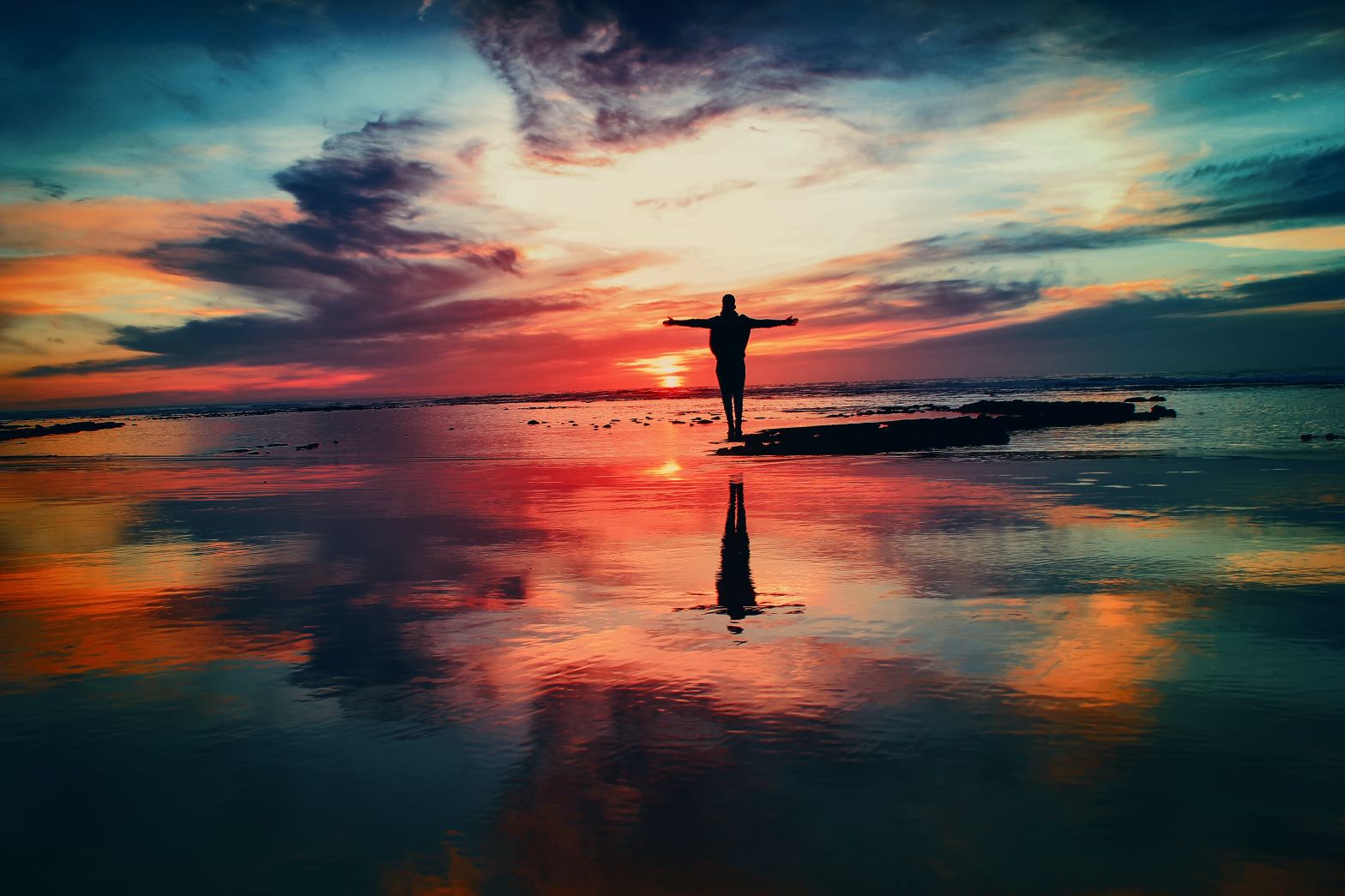 Silhouette of a person standing on a rock in the middle of water with arms stretched to the side with a sunrise on the horizon, the sky a brilliant mix of dark and light and reflected in the water below.